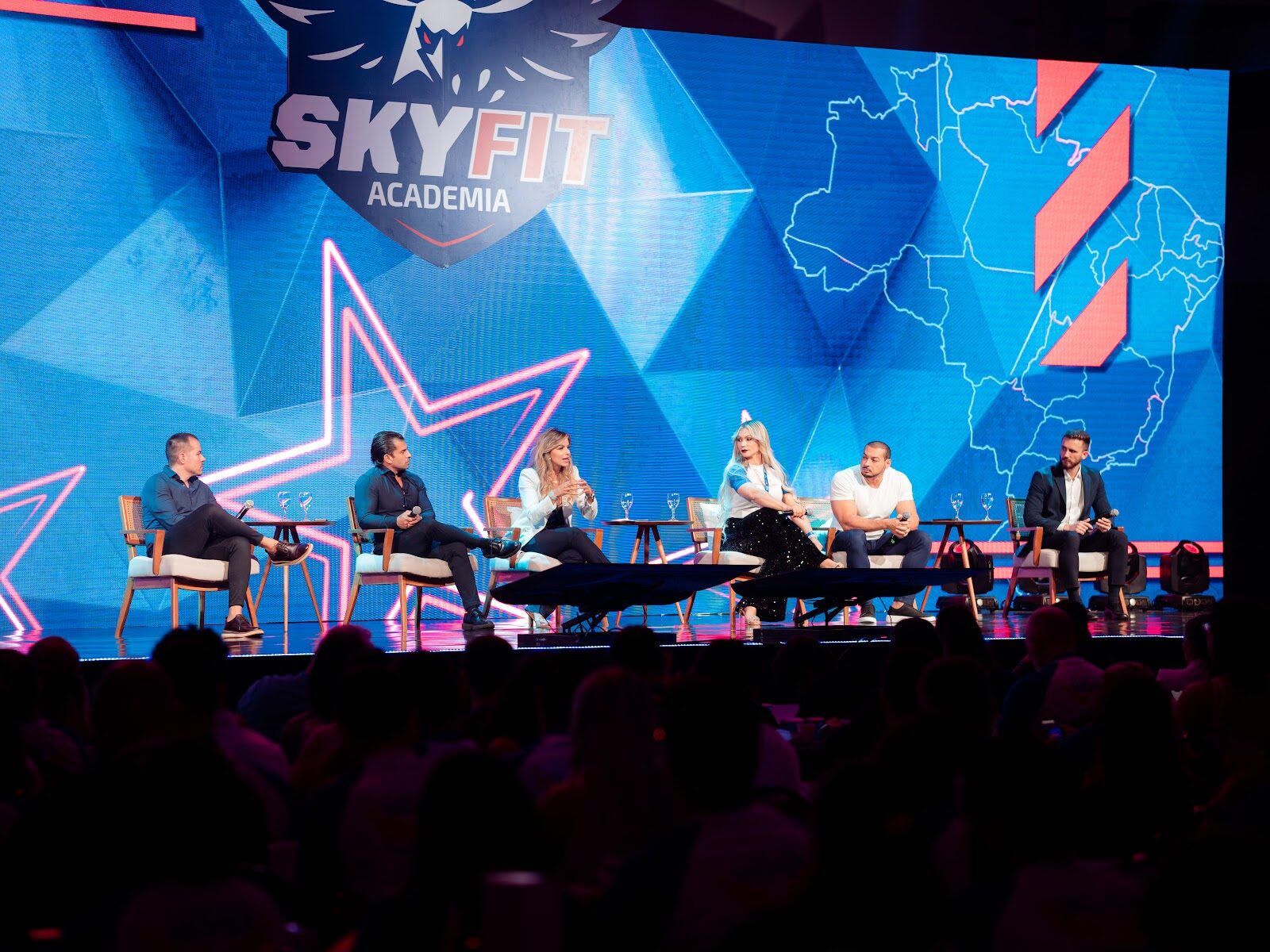 Mesa-redonda com seis painelistas em palco de convenção corporativa com painel LED ao fundo