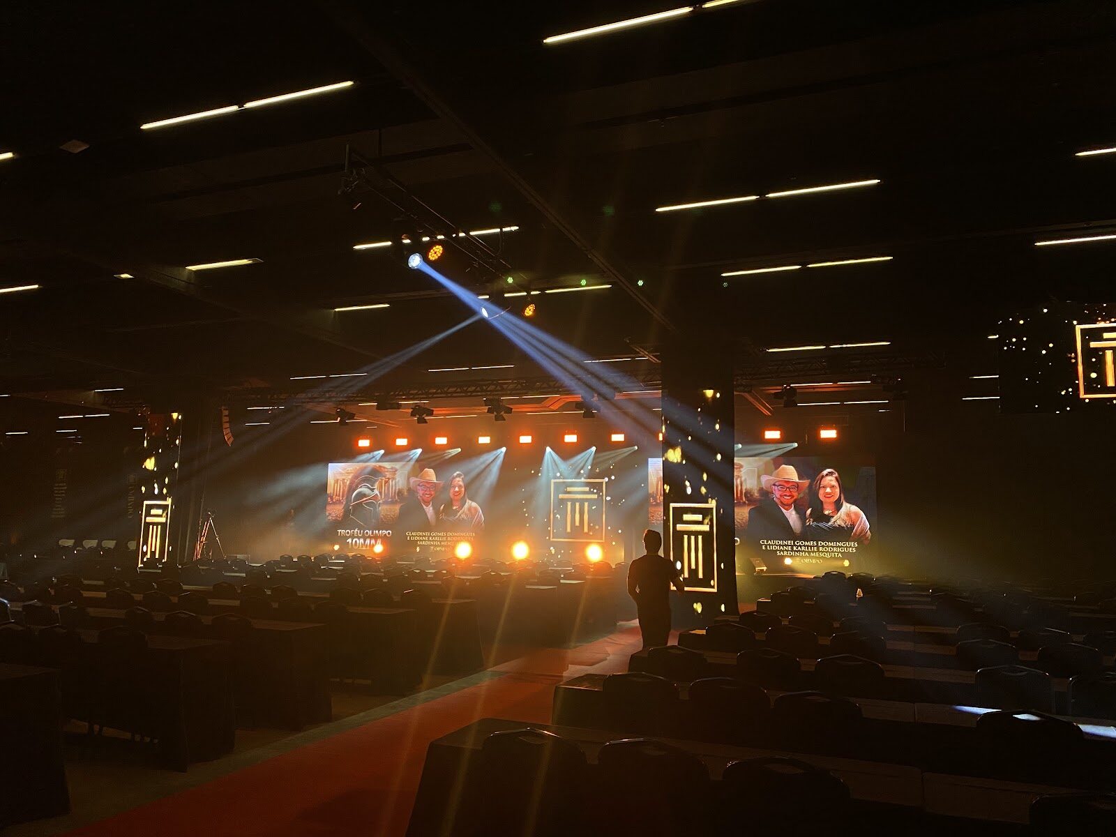 Palco de premiação corporativa com confete dourado e feixes de luz azuis