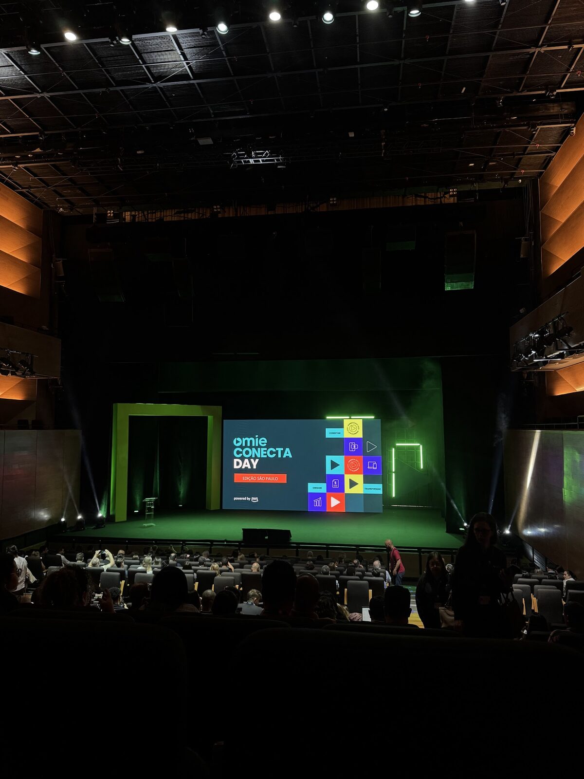 Plateia em auditório assistindo palco com telão LED widescreen verde do Omie Conecta Day