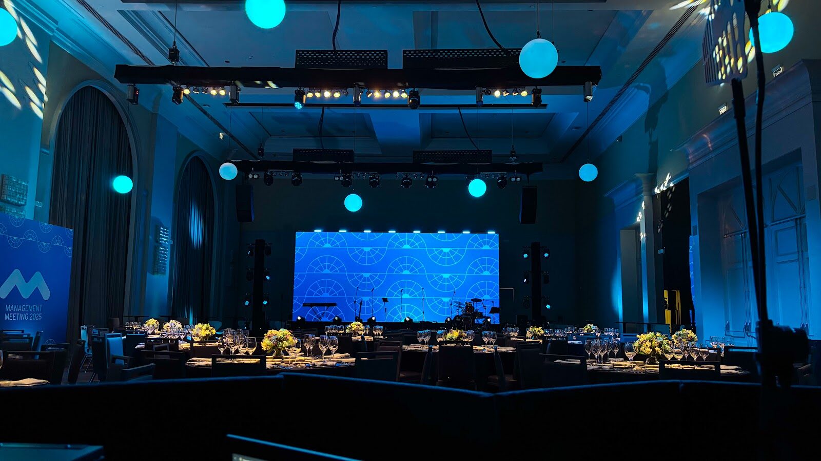 Salão de evento corporativo com painel LED azul no palco e esferas luminosas suspensas