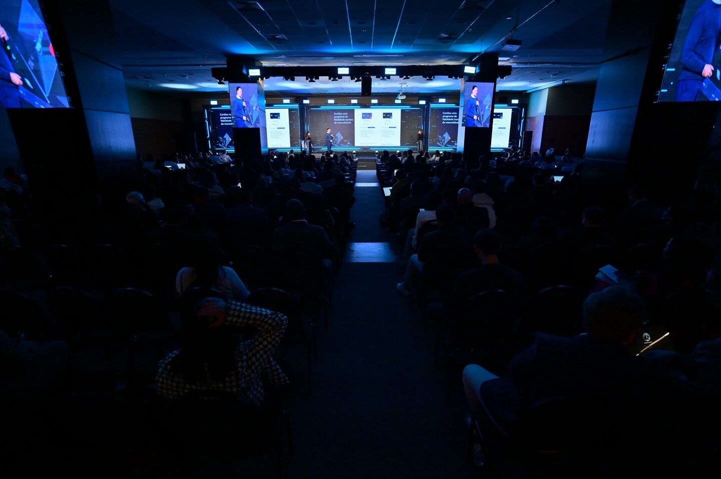 Auditório lotado em convenção corporativa com painel de LED central e telões laterais acima da plateia